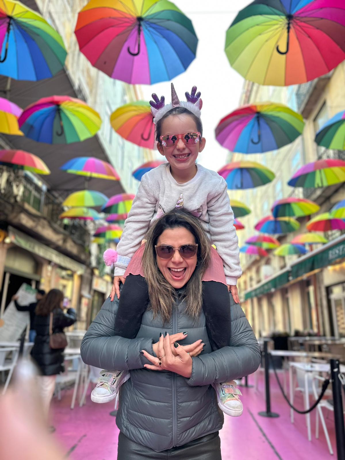 Mari carrying a smiling child on her shoulders beneath colorful rainbow umbrellas